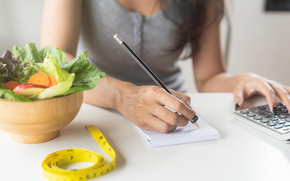 A woman sitting at a table next to a bowl of salad and a fitness tape measure using a calculator and writing results on a notepad