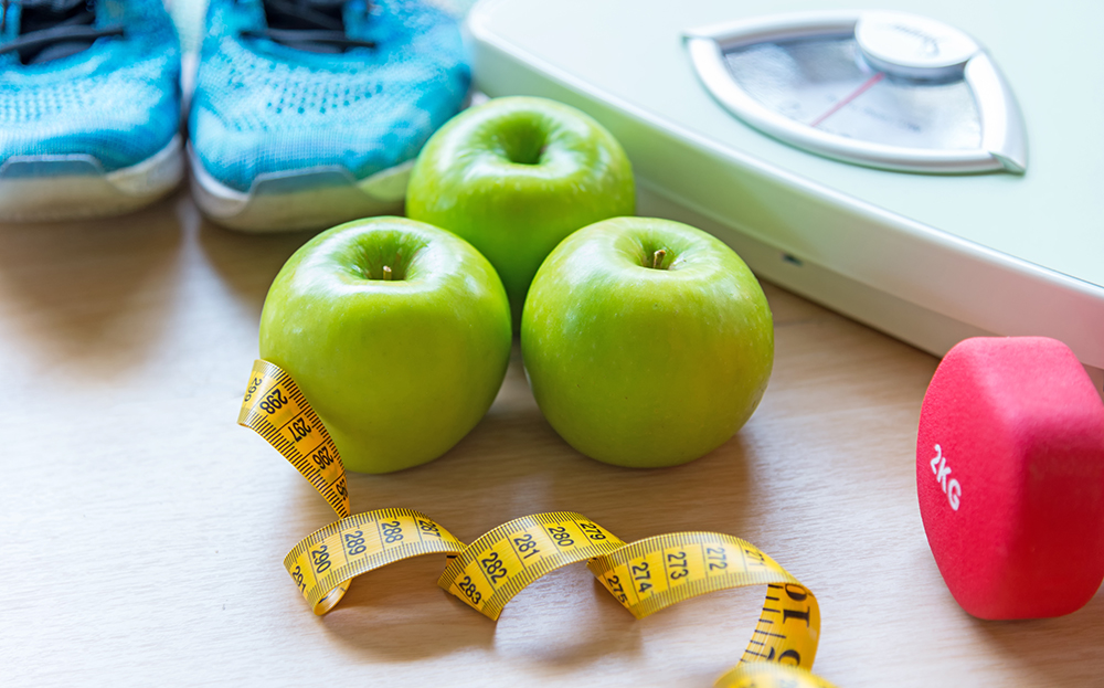 Apples, a pair of running shoes, a tape measure, and a weight on the floor next to a scale