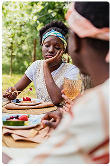 Two women sitting at a table full of food outdoors with one woman looking really bored while eating