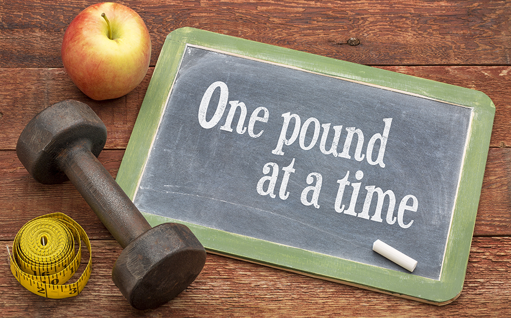An apple, dumbbell, tape measure, and chalkboard, with text one pound at a time, on a wooden table